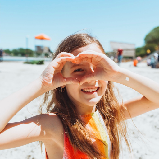 Smiling girl making a heart shape on a sunny beach, a UV-detecting sunscreen sticker on her wrist.