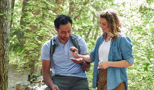 Two adults examining a small bottle of insect repellent while resting on a wooded trail surrounded by green foliage.