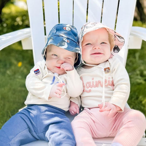 Two babies sitting on a white outdoor chair wearing sun hats and NatPat BuzzPatch stickers on their sleeves.