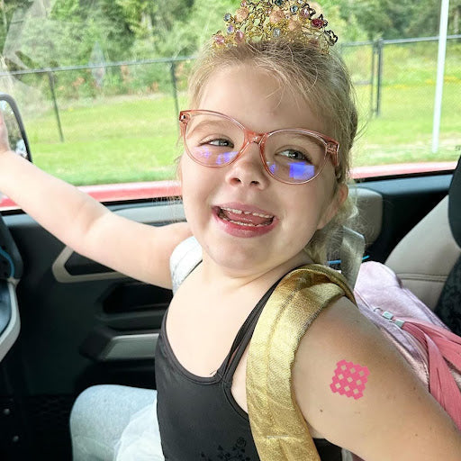 Smiling young girl wearing glasses and a crown with a pink NatPat MagicPatch Itch Relief Patch on her arm.