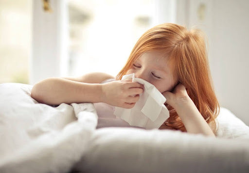 Young red-haired girl lying in bed blowing her nose into a tissue during allergy season.