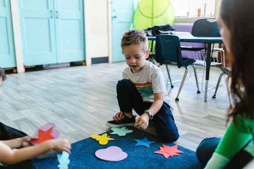 Young boy kneeling on a classroom floor sorting colorful paper shapes during a group learning activity.