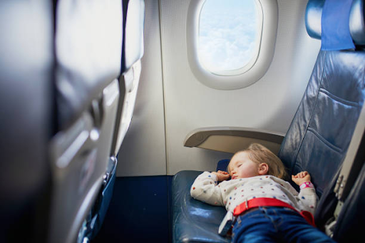 Toddler sleeping peacefully across airplane seats next to a sunlit cabin window.