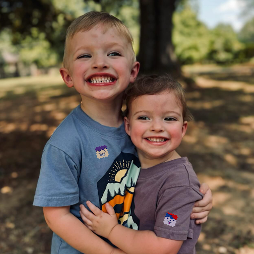 Two smiling children outdoors wearing mosquito repellent stickers on their t-shirts for mess-free protection.