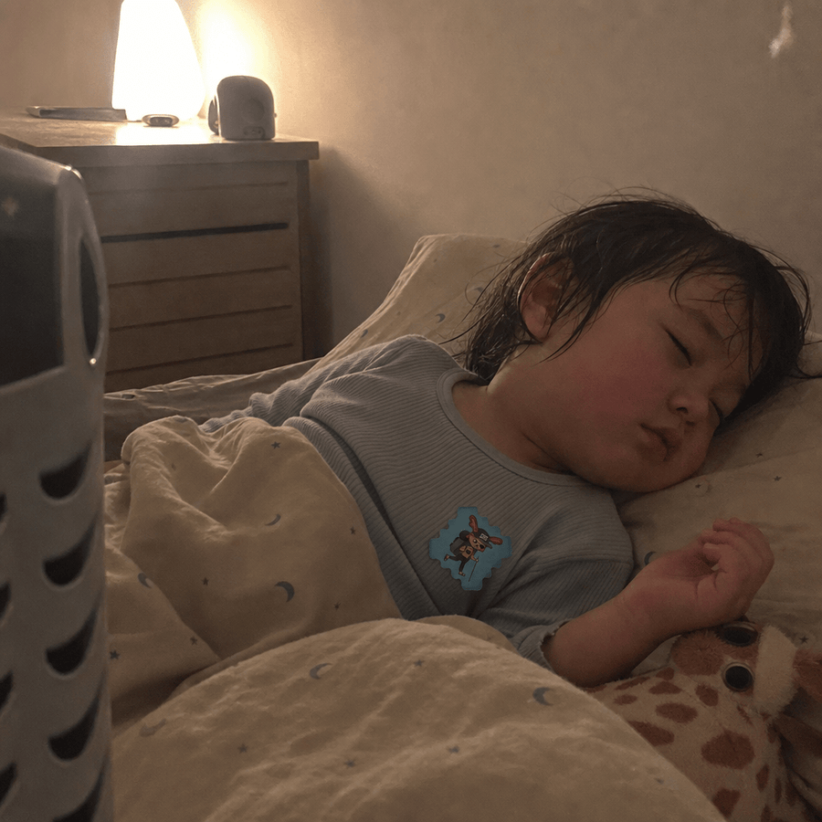 A parent leaning over a crib to place a StuffyPatch sticker on a sleeping child's pajama collar, warm bedside lamp light.