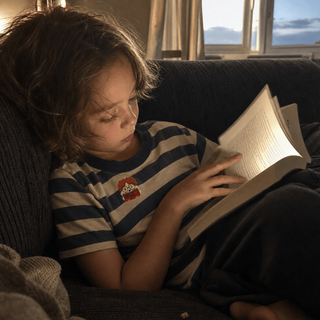 A young child in a navy-and-cream striped T-shirt with a NatPat sticker on the chest reads a book on a dark sofa by a window at dusk.