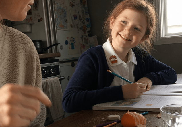 A smiling primary-school girl in a navy cardigan and white polo shirt, a NatPat sticker on her collar, writes in a workbook at a kitchen table while a parent stands beside her.