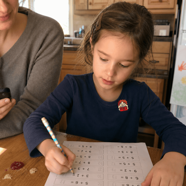 A girl in a navy top works through a page of addition sums at a kitchen table, a FocusPatch sticker on her chest, with a parent sitting beside her.