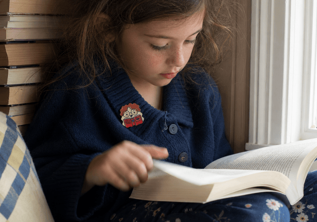 A girl in a navy cardigan reads a thick book by a window, a FocusPatch sticker on her chest and a stack of books beside her.