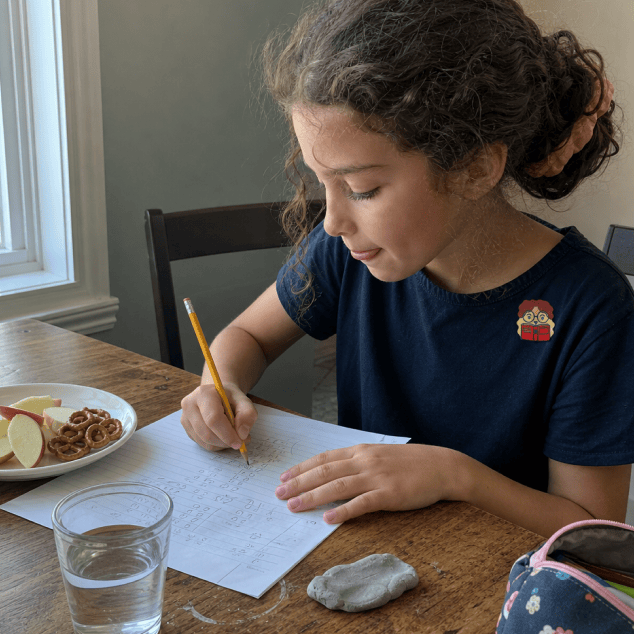 A girl in a navy T-shirt with a FocusPatch sticker on her chest writes on a lined worksheet at a wooden kitchen table, a plate of apple slices and pretzels and a glass of water beside her.