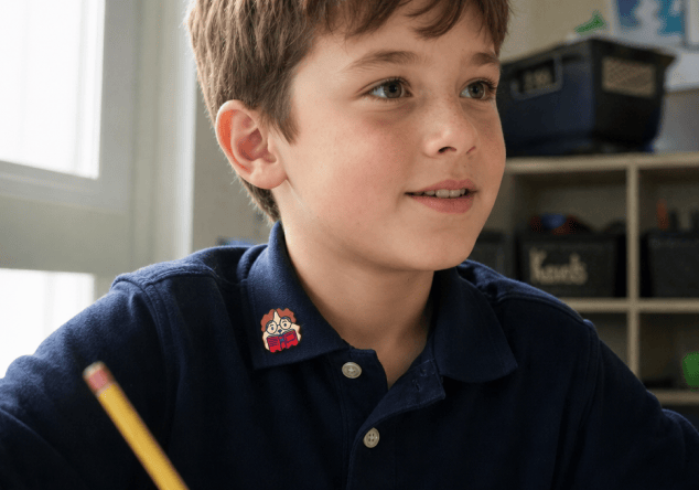 A boy in a navy polo shirt with a FocusPatch sticker on his collar looks up smiling in a classroom lined with shelves of storage bins, a pencil in his hand.