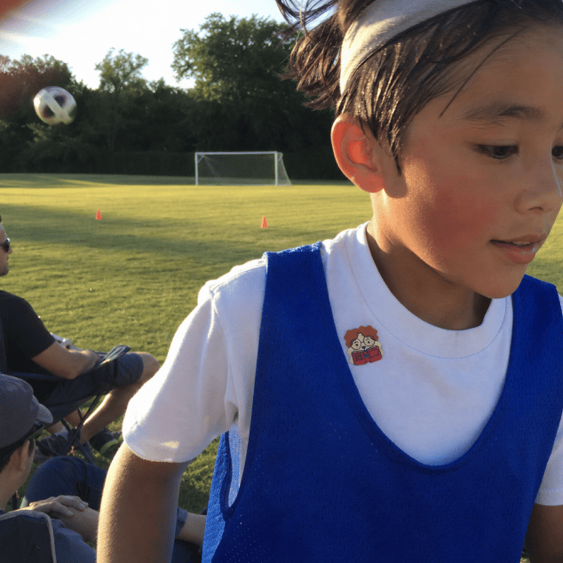 A young footballer in a white tee and blue training bib on a grass pitch at golden hour, a FocusPatch sticker on the shirt near the collar and a ball in the air behind.