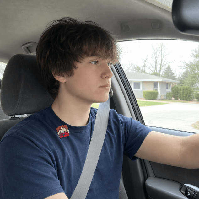 A teenage boy in a navy t-shirt with a FocusPatch sticker on his chest sits belted in the driver's seat, hand on the wheel, looking ahead through the windscreen at a suburban street.
