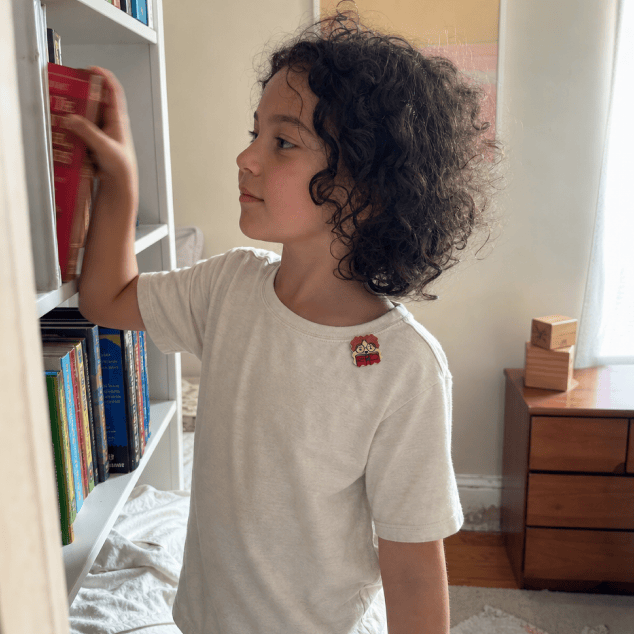 A child with curly dark hair in a cream t-shirt with a FocusPatch sticker on the chest reaches up to pull a red book from a white bookshelf in a bedroom.