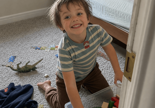 A smiling young boy in a blue-and-cream striped t-shirt with a FocusPatch sticker on his chest kneels on a bedroom carpet beside a toy crocodile and scattered puzzle pieces, pulling a clear storage box out from under the bed.
