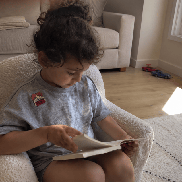 A young child sits curled in a cream bouclé armchair reading an open book, a cartoon FocusPatch sticker on the chest of their grey T-shirt.