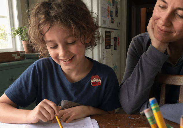A smiling child in a navy T-shirt with a FocusPatch sticker on the chest works through a worksheet at a kitchen table while a parent leans on one hand and watches.