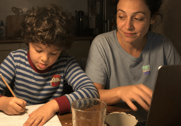 A curly-haired child writes with a pencil at a kitchen table in the evening while their mother works on a laptop beside them, each wearing a NatPat sticker on their top.