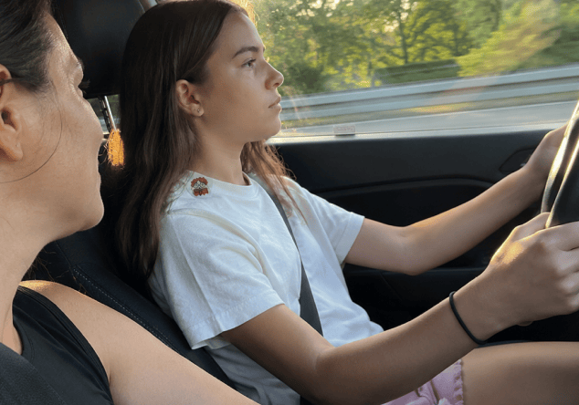 A teenage girl in a white t-shirt with a FocusPatch sticker on her shoulder drives with both hands on the wheel while a woman sits watching from the passenger seat, trees blurring past the window.