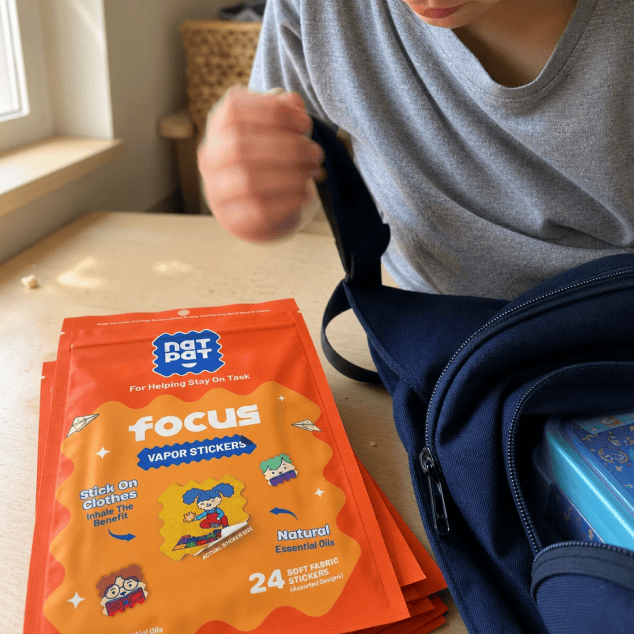 A child in a grey t-shirt sits at a kitchen table holding the strap of a navy backpack, with a stack of orange NATPAT Focus vapor sticker pouches on the table and a lunchbox in the open bag.