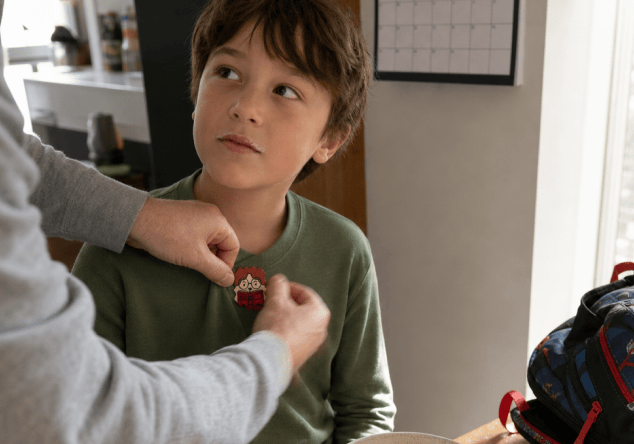 A parent presses a cartoon Focus sticker onto the chest of a boy's green shirt at the breakfast table, with a cereal bowl and a packed backpack beside them.