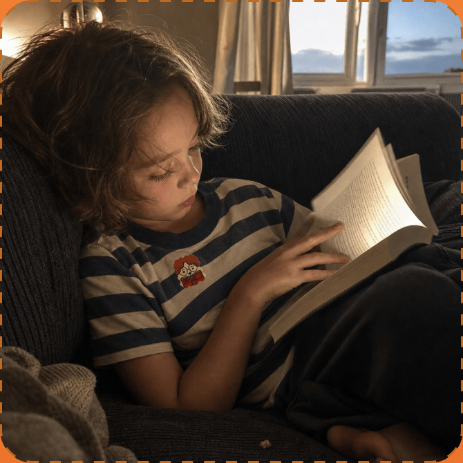 A young child in a navy-and-cream striped T-shirt with a NatPat sticker on the chest reads a book on a dark sofa by a window at dusk.