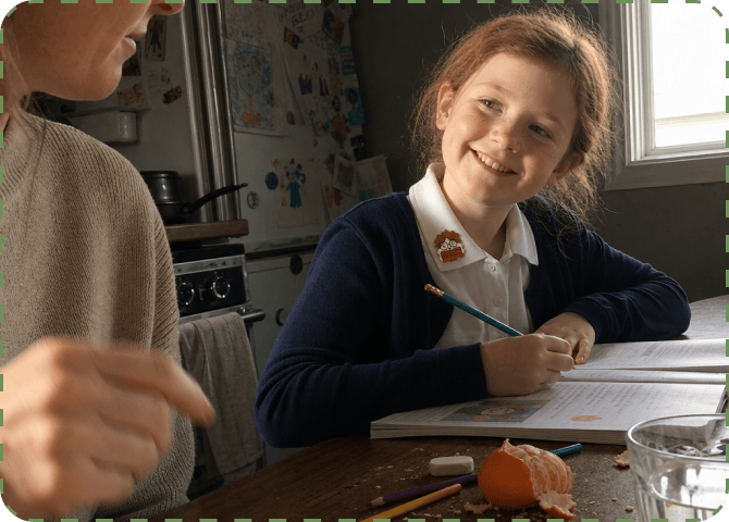 A smiling primary-school girl in a navy cardigan and white polo shirt, a NatPat sticker on her collar, writes in a workbook at a kitchen table while a parent stands beside her.