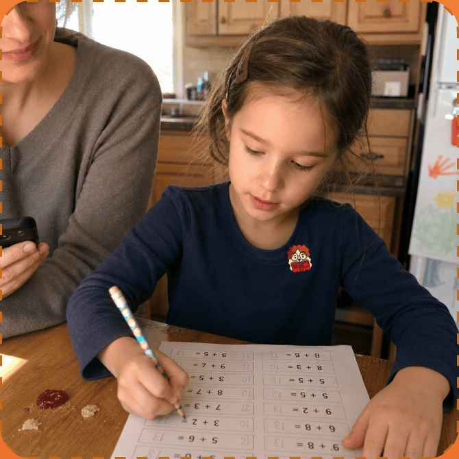A girl in a navy top works through a page of addition sums at a kitchen table, a FocusPatch sticker on her chest, with a parent sitting beside her.