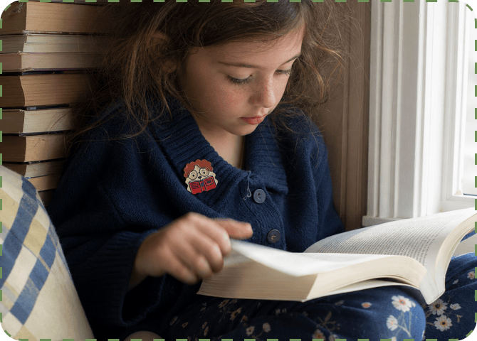 A girl in a navy cardigan reads a thick book by a window, a FocusPatch sticker on her chest and a stack of books beside her.