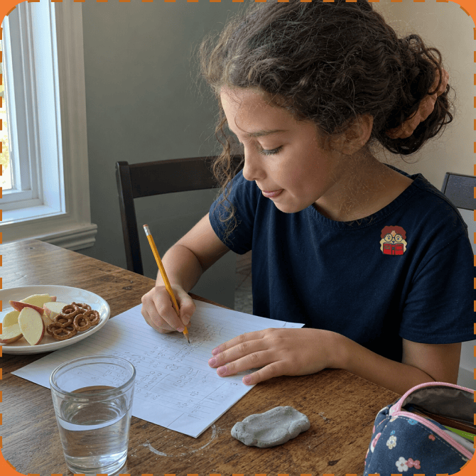 A girl in a navy T-shirt with a FocusPatch sticker on her chest writes on a lined worksheet at a wooden kitchen table, a plate of apple slices and pretzels and a glass of water beside her.