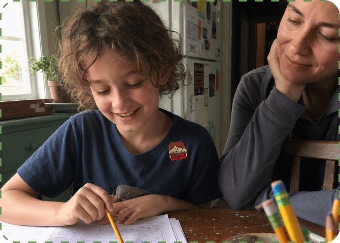 A smiling child in a navy T-shirt with a FocusPatch sticker on the chest works through a worksheet at a kitchen table while a parent leans on one hand and watches.