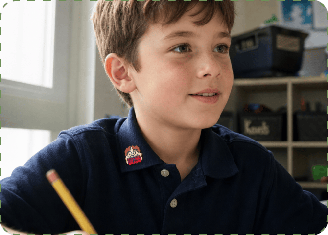 A boy in a navy polo shirt with a FocusPatch sticker on his collar looks up smiling in a classroom lined with shelves of storage bins, a pencil in his hand.