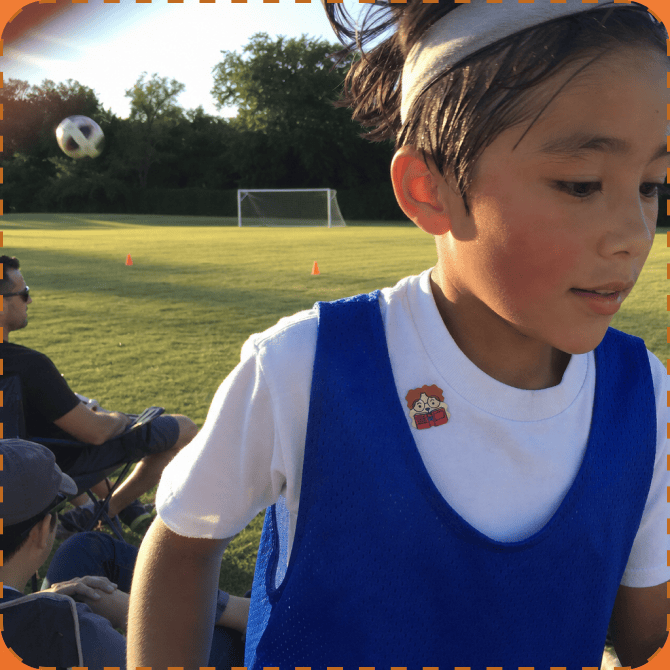 A young footballer in a white tee and blue training bib on a grass pitch at golden hour, a FocusPatch sticker on the shirt near the collar and a ball in the air behind.