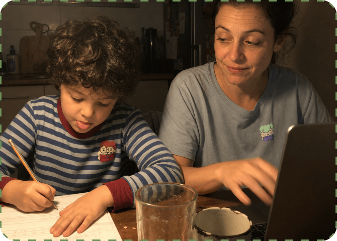 A curly-haired child writes with a pencil at a kitchen table in the evening while their mother works on a laptop beside them, each wearing a NatPat sticker on their top.