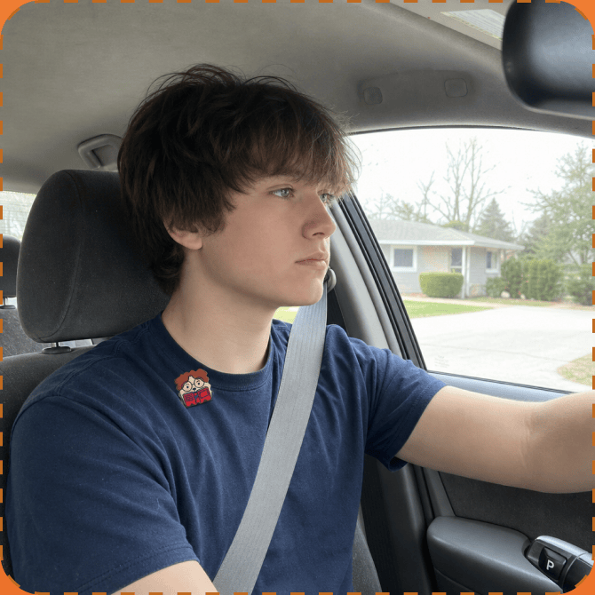 A teenage boy in a navy t-shirt with a FocusPatch sticker on his chest sits belted in the driver's seat, hand on the wheel, looking ahead through the windscreen at a suburban street.