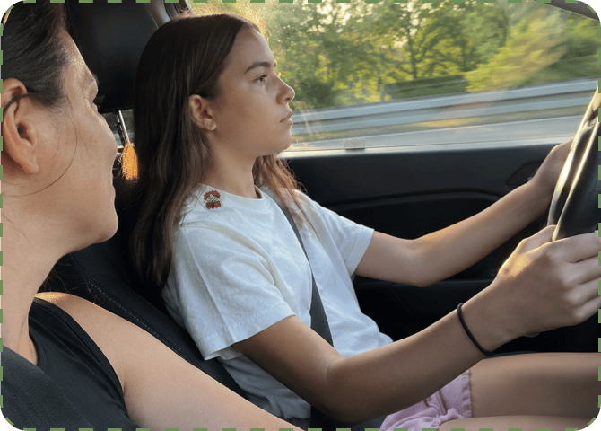 A teenage girl in a white t-shirt with a FocusPatch sticker on her shoulder drives with both hands on the wheel while a woman sits watching from the passenger seat, trees blurring past the window.