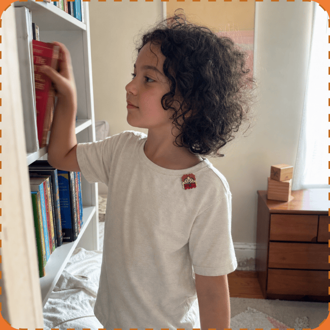 A child with curly dark hair in a cream t-shirt with a FocusPatch sticker on the chest reaches up to pull a red book from a white bookshelf in a bedroom.