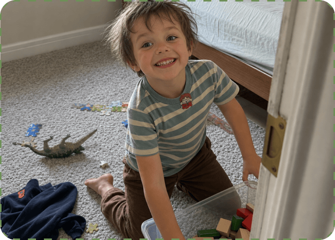 A smiling young boy in a blue-and-cream striped t-shirt with a FocusPatch sticker on his chest kneels on a bedroom carpet beside a toy crocodile and scattered puzzle pieces, pulling a clear storage box out from under the bed.