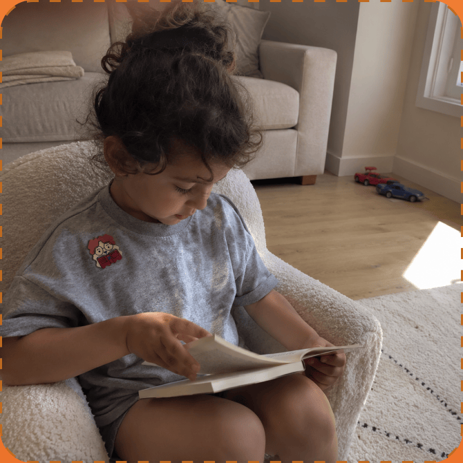 A young child sits curled in a cream bouclé armchair reading an open book, a cartoon FocusPatch sticker on the chest of their grey T-shirt.
