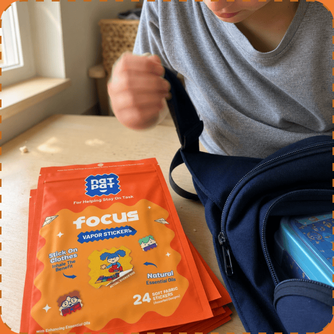 A child in a grey t-shirt sits at a kitchen table holding the strap of a navy backpack, with a stack of orange NATPAT Focus vapor sticker pouches on the table and a lunchbox in the open bag.