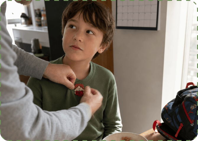 A parent presses a cartoon Focus sticker onto the chest of a boy's green shirt at the breakfast table, with a cereal bowl and a packed backpack beside them.