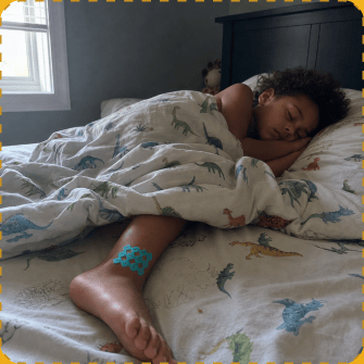 A young child asleep in bed under a dinosaur-print duvet, with a blue MagicPatch stuck on their lower leg.