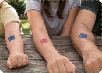 A child, a woman and a man rest their forearms side by side on a wooden picnic table, each wearing a grid-shaped MagicPatch on their arm.
