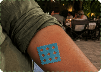 Close-up of an adult’s upper arm at an outdoor restaurant table with a blue MagicPatch grid sticker pressed onto the skin.