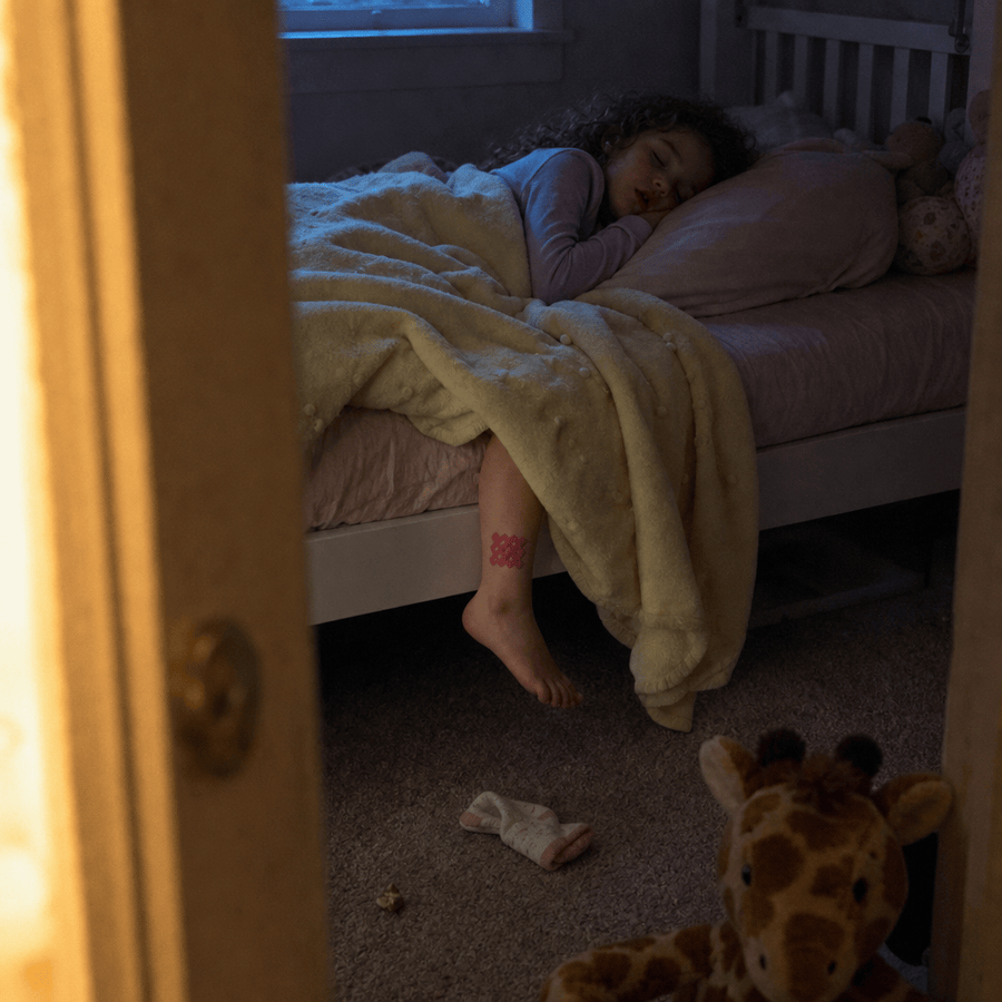A young girl asleep in bed at night, seen through a half-open bedroom door, with a red grid patch on her calf and a plush giraffe toy on the carpet below.