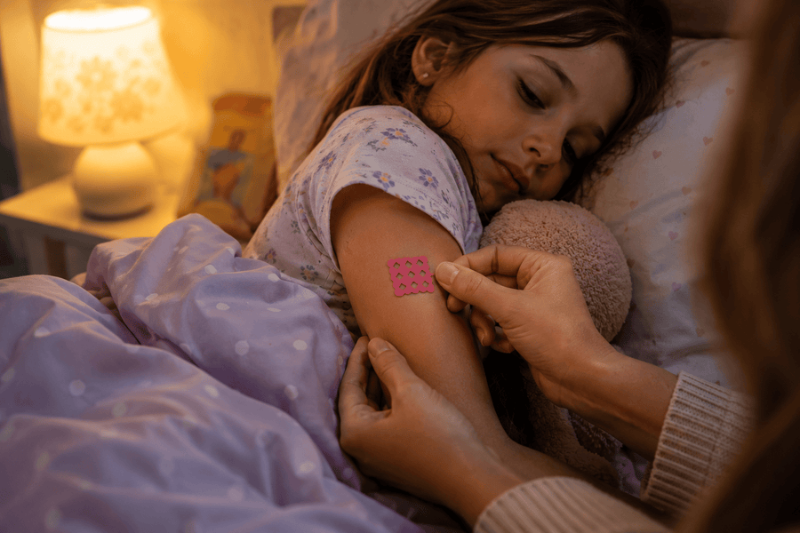 A parent in warm lamplight pressing a pink grid patch onto the upper arm of a sleepy child tucked under a lilac blanket.