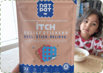 A hand holds up a tan NatPat Itch Relief Stickers pouch at a kitchen table while a young girl in a strawberry-print top sits behind it with a bowl of cereal.