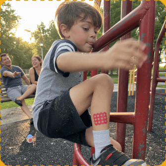 A young boy on a red playground climbing frame at golden hour has a red grid-patterned MagicPatch stuck on his shin, while two smiling parents look on from a bench behind him.