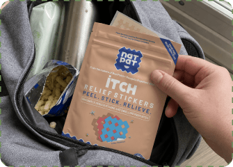 A hand pulls a tan NatPat Itch Relief Stickers pouch out of an open backpack pocket, next to a spilling bag of puffed corn snacks.