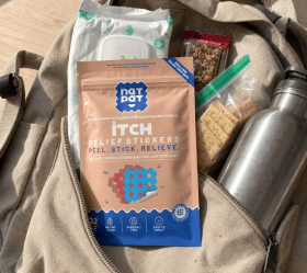 A NatPat Itch Relief Stickers pouch peeking out of an open backpack pocket, next to a granola bar, a pack of crackers, a first-aid roll and a metal water bottle.